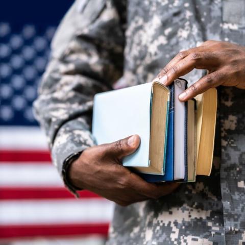 A uniformed servicemember in front of a flag carrying books