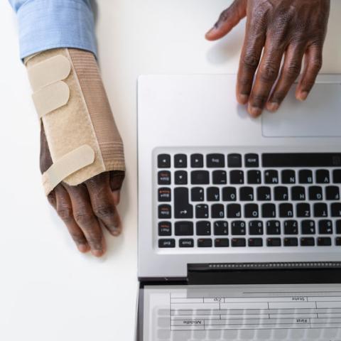 An office worker's hands on a laptop. One hand has a brace.